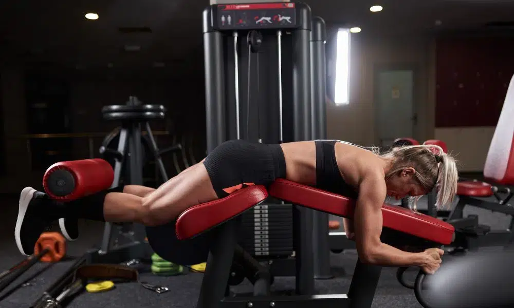 mujer rubia haciendo leg curl tumbada en gimnasio oscuro