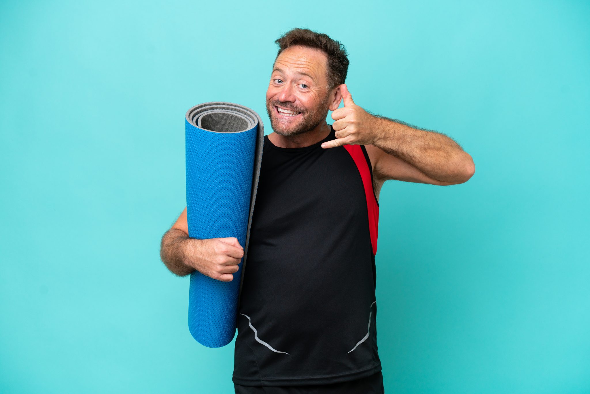 middle age sport man going to yoga classes while holding a mat isolated on blue background making phone gesture. call me back sign